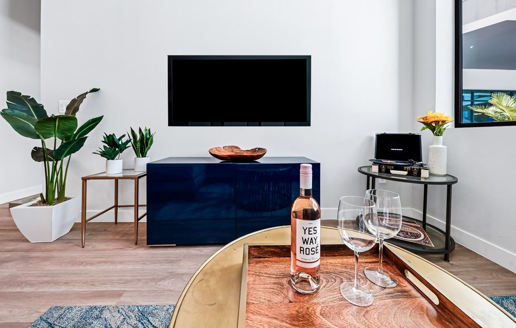 A living room with a blue couch, a wooden table with a bottle of wine and glasses, and a TV mounted on the wall.
