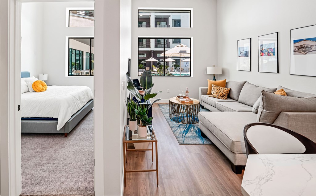 A modern living room with a grey sofa, a bed with white bedding, a wooden coffee table, and a large window with a view of the city.