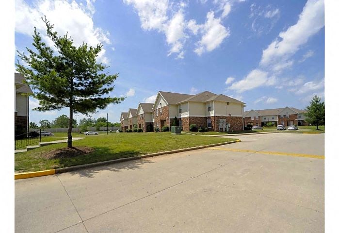 Exterior of Woodhaven Park apartment buildings with parking spots, grass areas, and trees during the daytime.