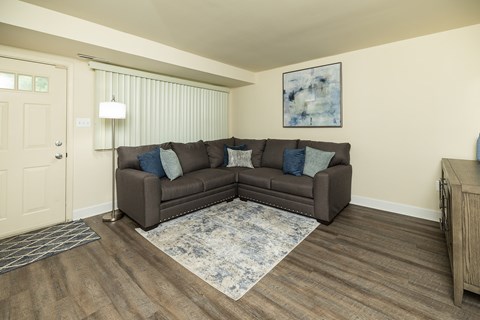 A brown couch with blue pillows sits in a room with wooden floors and a white door at The Orchards at Severn Townhomes*, Severn, MD 21144.