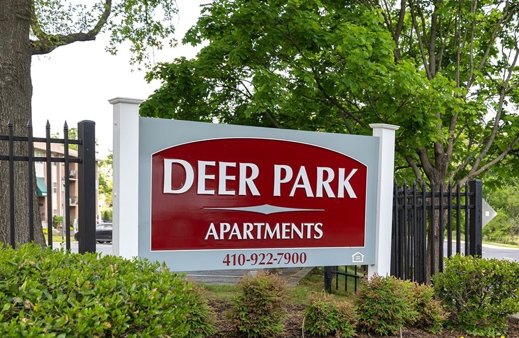 A sign for Deer Park Apartments is displayed in front of a fence and greenery.