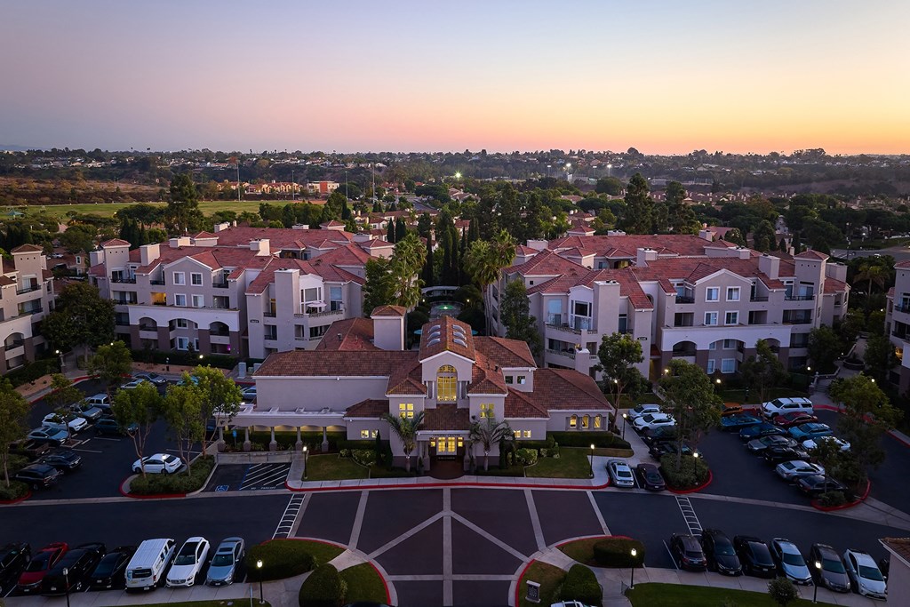 A sunset view of a large white building with a red roof and a parking lot in front at Valentia Apartments in La Jolla.