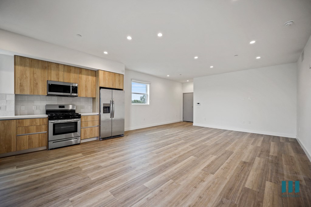 an empty living room with wood flooring and a kitchen with stainless steel appliances