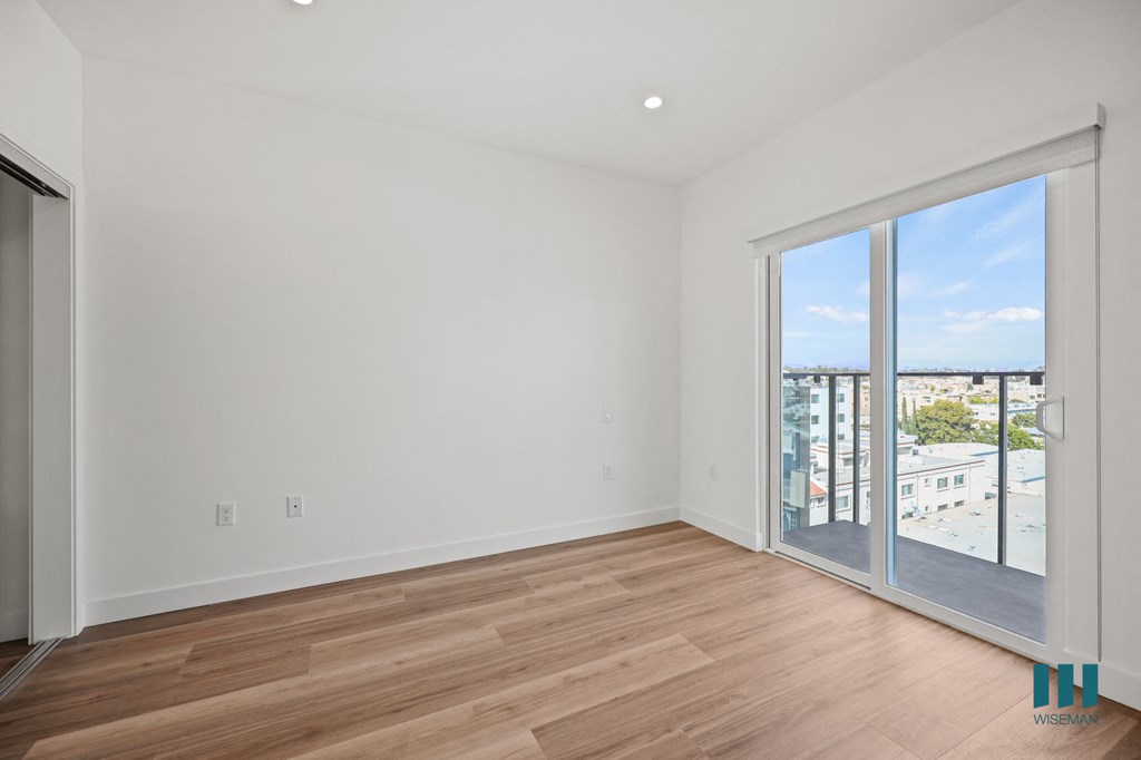 A room with a sliding glass door leading to a balcony with a view of buildings and a blue sky.
