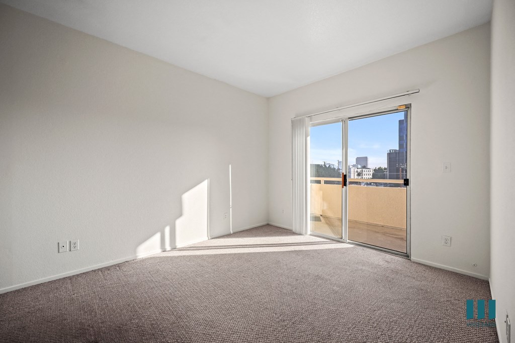 A large empty room with a carpeted floor and a view of the city through a sliding glass door.