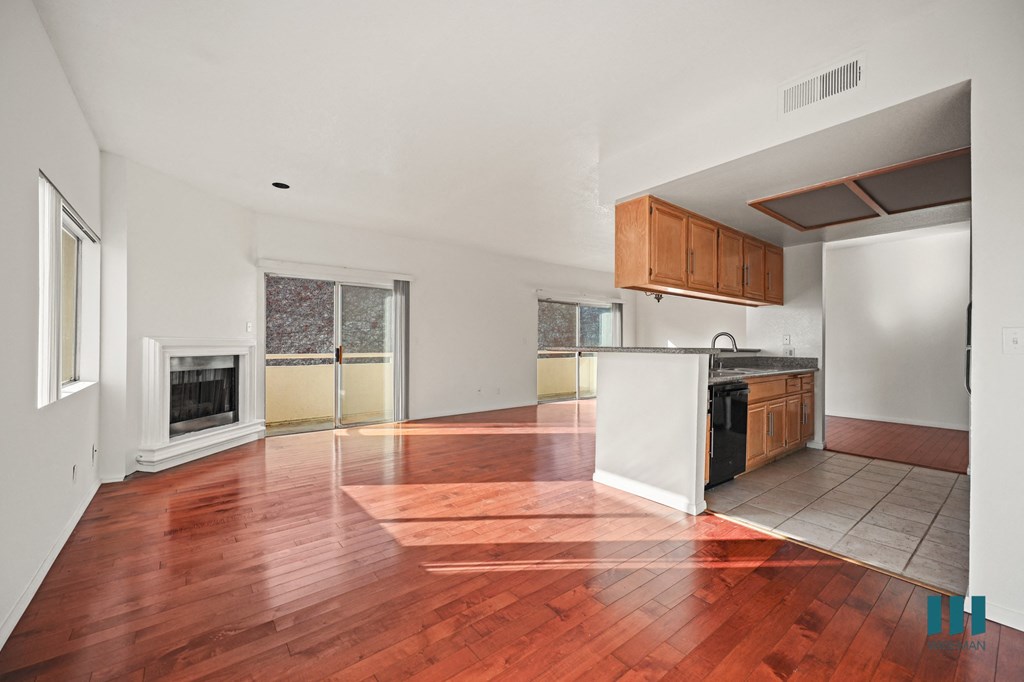 A large empty room with wood flooring and a kitchen area to the right.