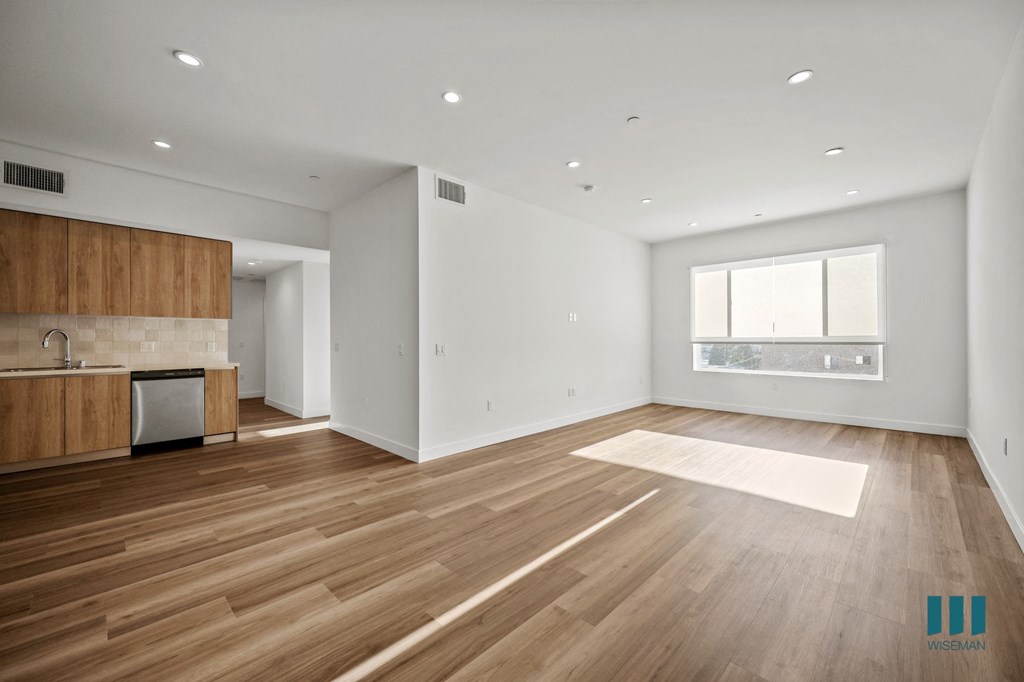 A spacious room with wooden flooring and a kitchen area with white walls.