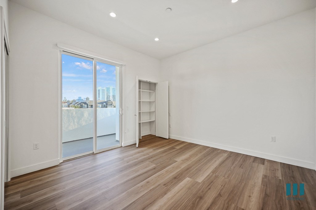 A room with a sliding glass door leading to a balcony, wooden flooring, and a white wall with a shelf.
