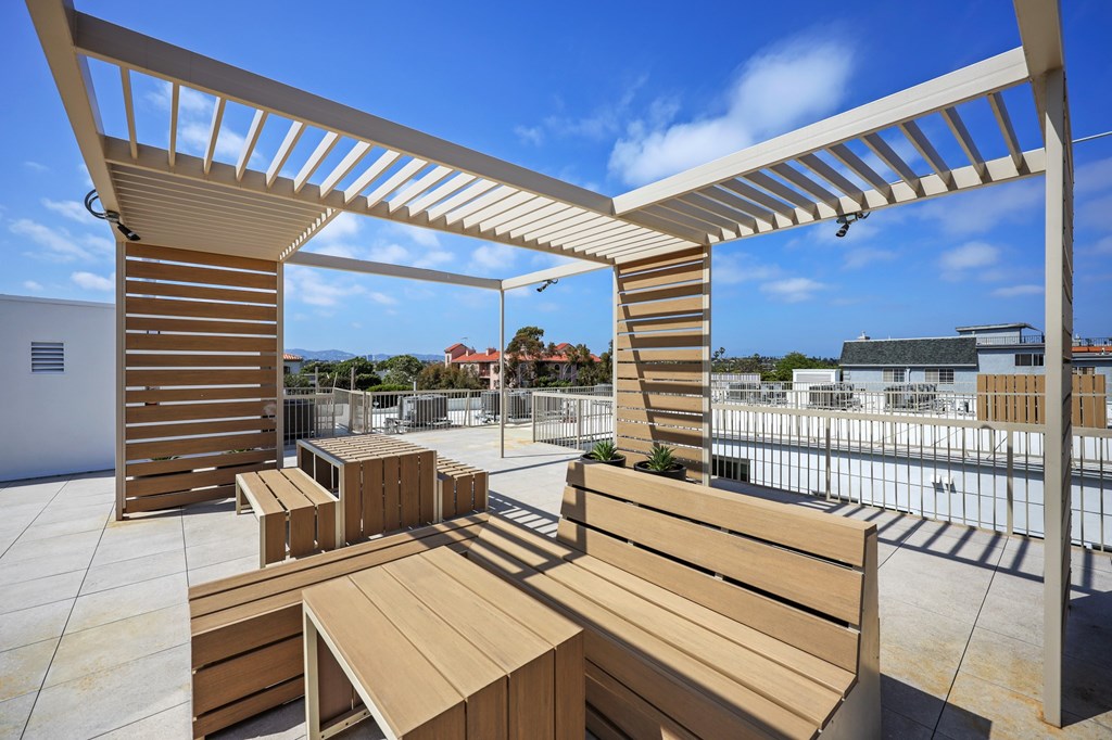 A wooden bench and table are under a white pergola on a rooftop.