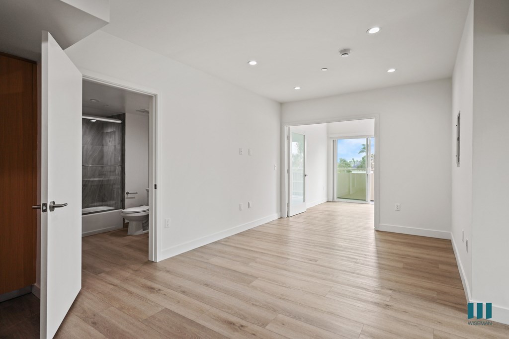 A room with wooden floors and white walls, with a door open to a bathroom.