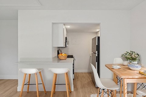 A kitchen with white walls and wooden floors, featuring a bar area with stools, a dining table with chairs, and a refrigerator.