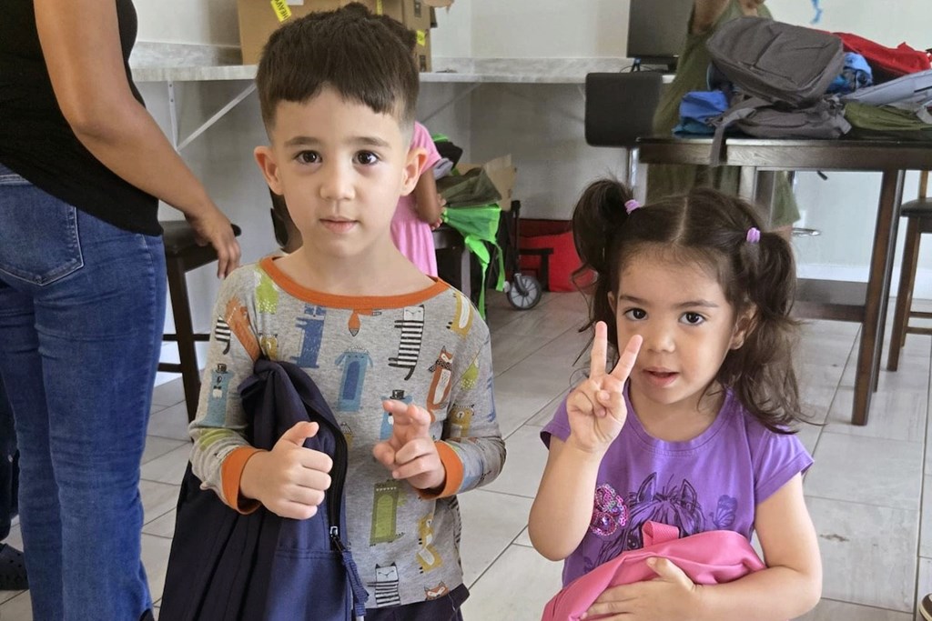 A young boy and girl are standing in a room with a table and chairs in the background.