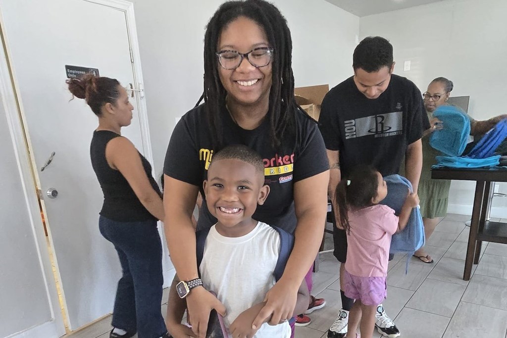 A woman with glasses and dreadlocks is standing in a room with a child in front of her.