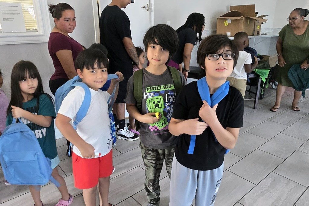 A group of children are standing in a room with a woman in the background.