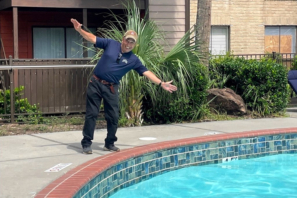 A man in a blue shirt and black pants is standing by a pool with his arms outstretched.