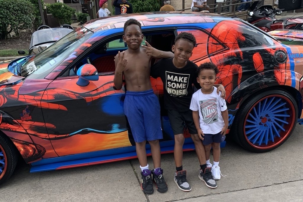 Two boys stand in front of a car with a black and orange design.