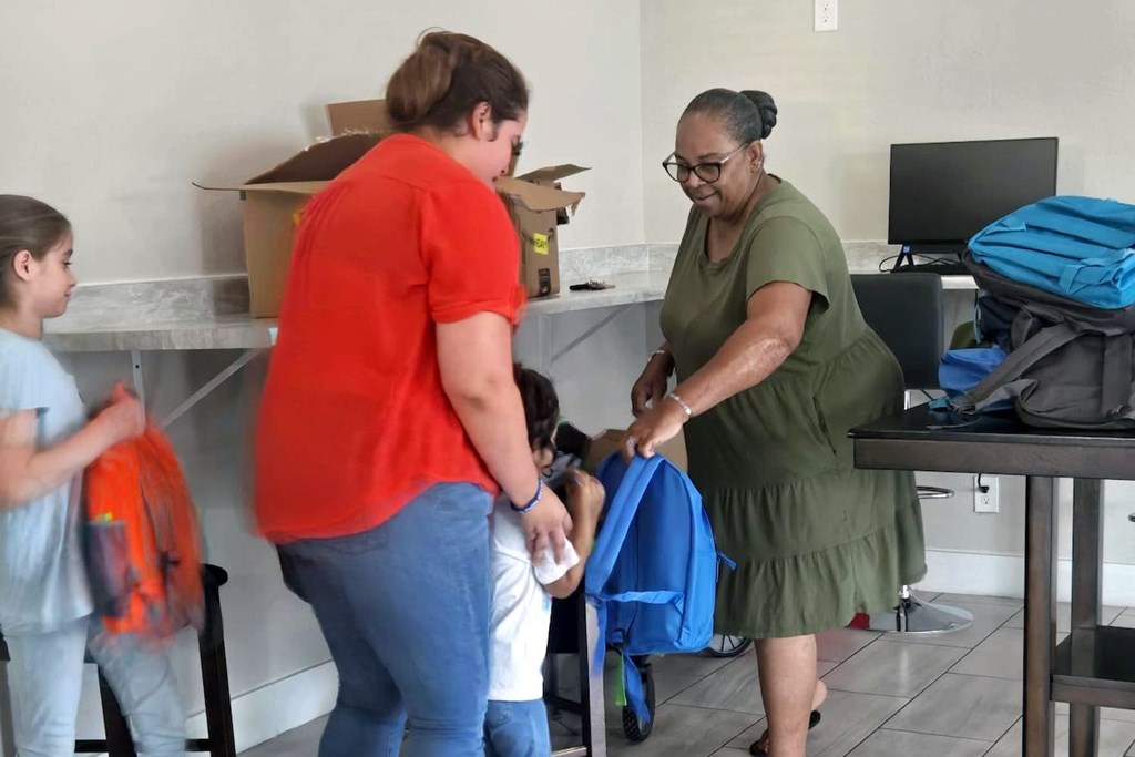 A woman in a green dress is helping a child with their backpack.