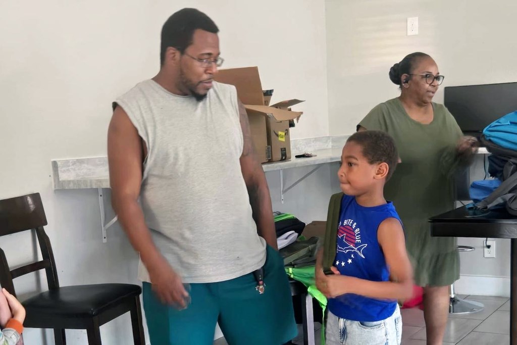 A man in a grey shirt and green pants is standing in a room with a woman in a green shirt and a boy in a blue shirt.