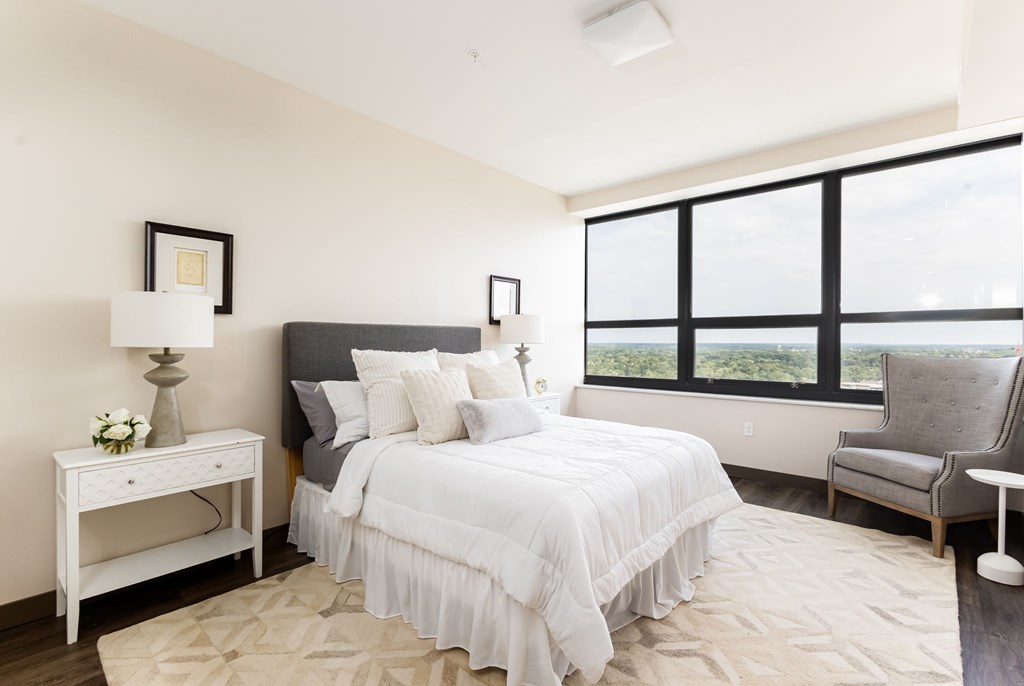 A bedroom with a large bed, a nightstand with a lamp, a chair, and a window with a view.