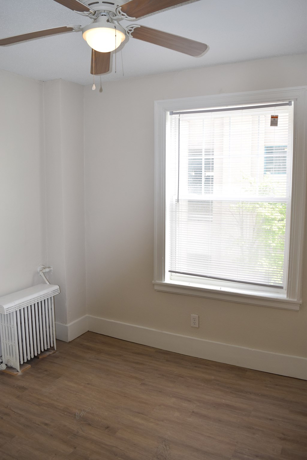 A room with a ceiling fan, a window with blinds, and a radiator on the floor.