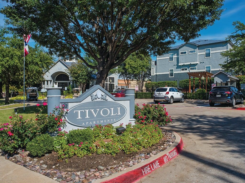 Apartments in North Dallas - Tivoli - Community Entrance with Gray Building, Entrance Sign, Lush Greenery, and Parking Lot