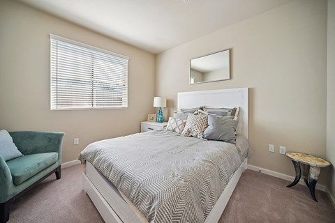 A bedroom with a bed, a green chair, a nightstand with a lamp, and a mirror on the wall.