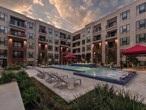 A large swimming pool is surrounded by lounge chairs and umbrellas in front of apartment buildings.