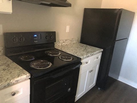 A black stove top oven with four burners and a digital display is in a kitchen with a black refrigerator and a marble counter top.