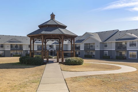 A gazebo is in the middle of a grassy area in front of apartment buildings.