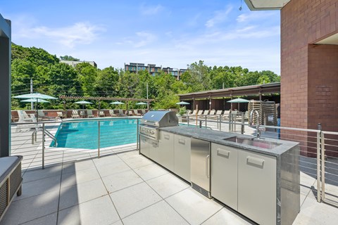 An outdoor grilling station with a BBQ and ample counter and cabinet space near a swimming pool surrounded by lounge chairs, umbrellas, and large trees.