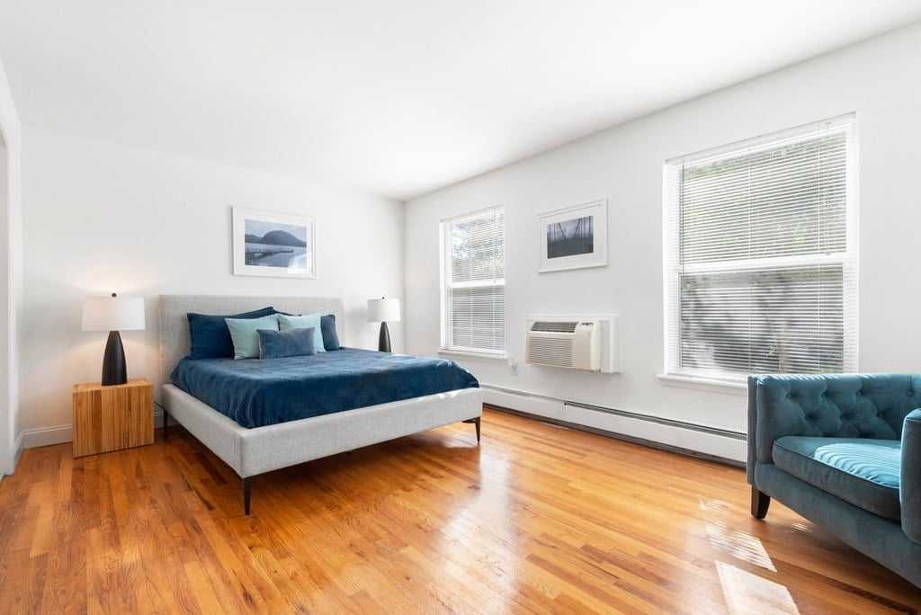 A bedroom with hardwood-style flooring, two large windows, and a portable cooling/heating unit on the wall.