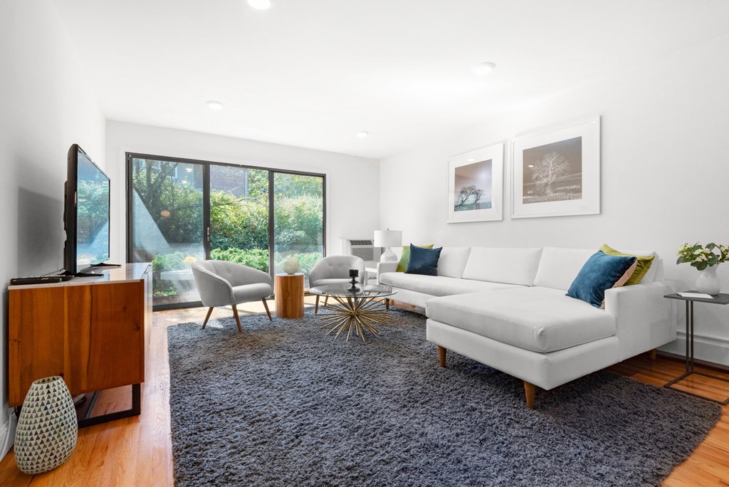 A modern living room with recessed lighting, hardwood-style flooring, and three large sliding doors to a private patio with lush greenery.