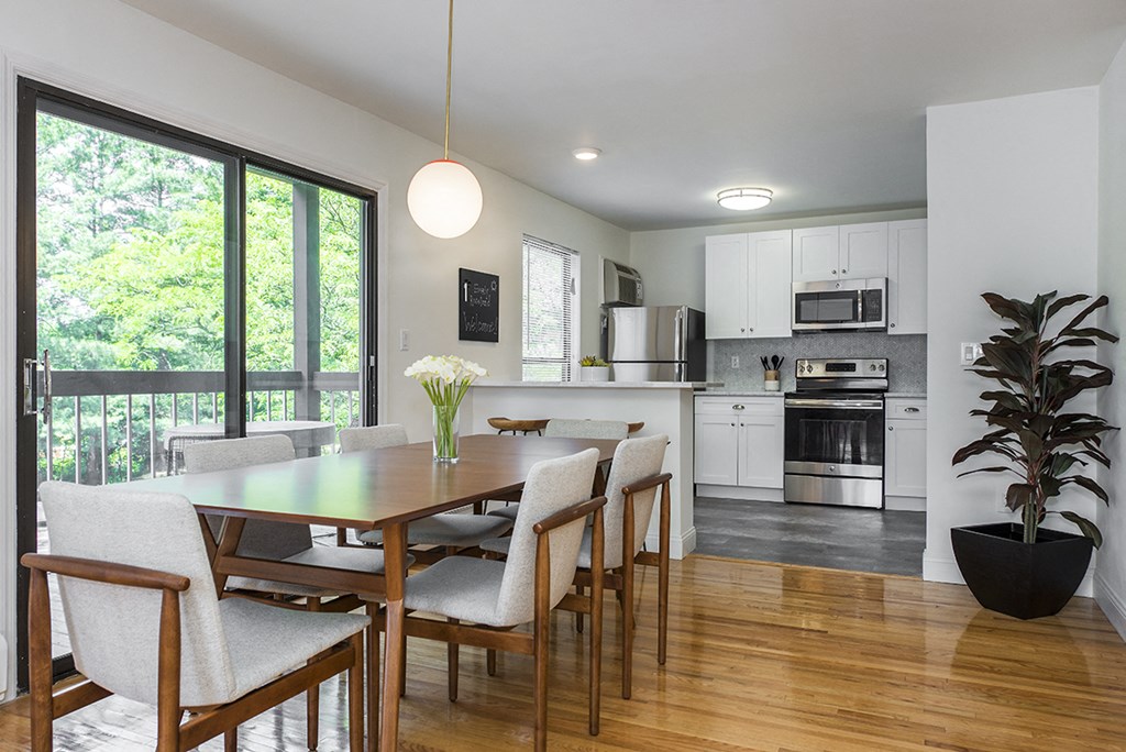 A dining room with hardwood-style flooring and a modern pendant light near a kitchen and double sliding doors leading to a private balcony.