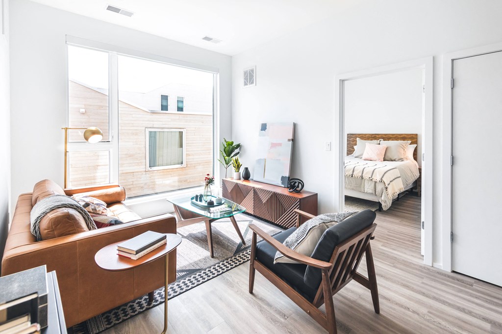 A living room with a brown sofa, a glass coffee table, and a bed in the background.