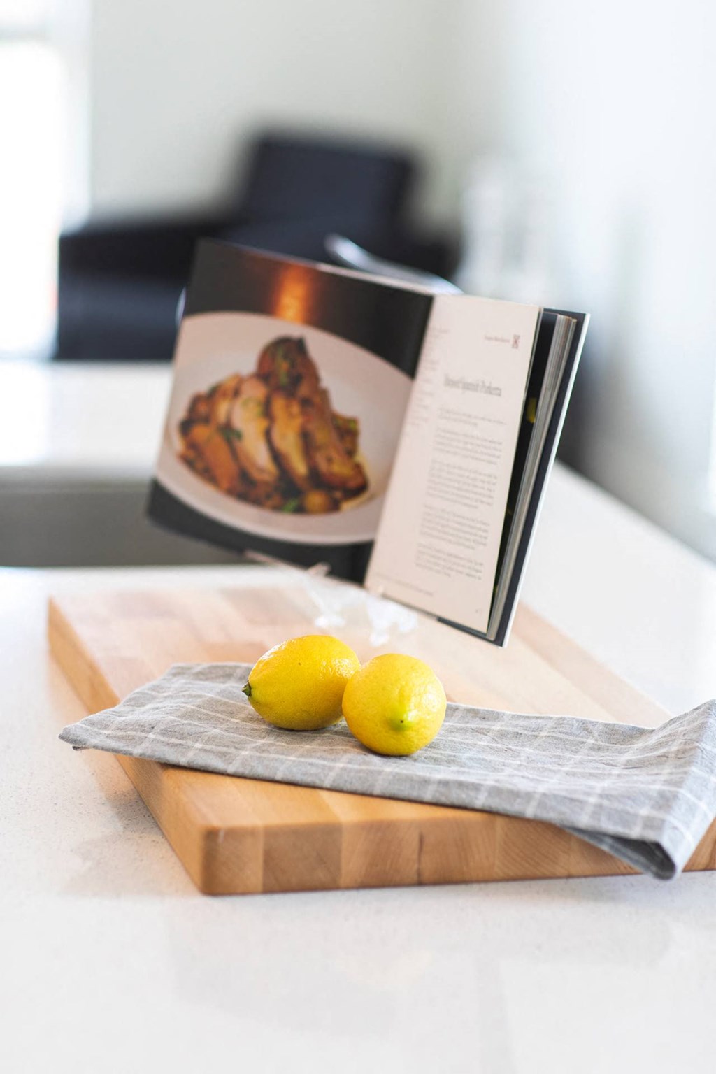 A book is open to a recipe for chicken, with two lemons on a cutting board beside it.