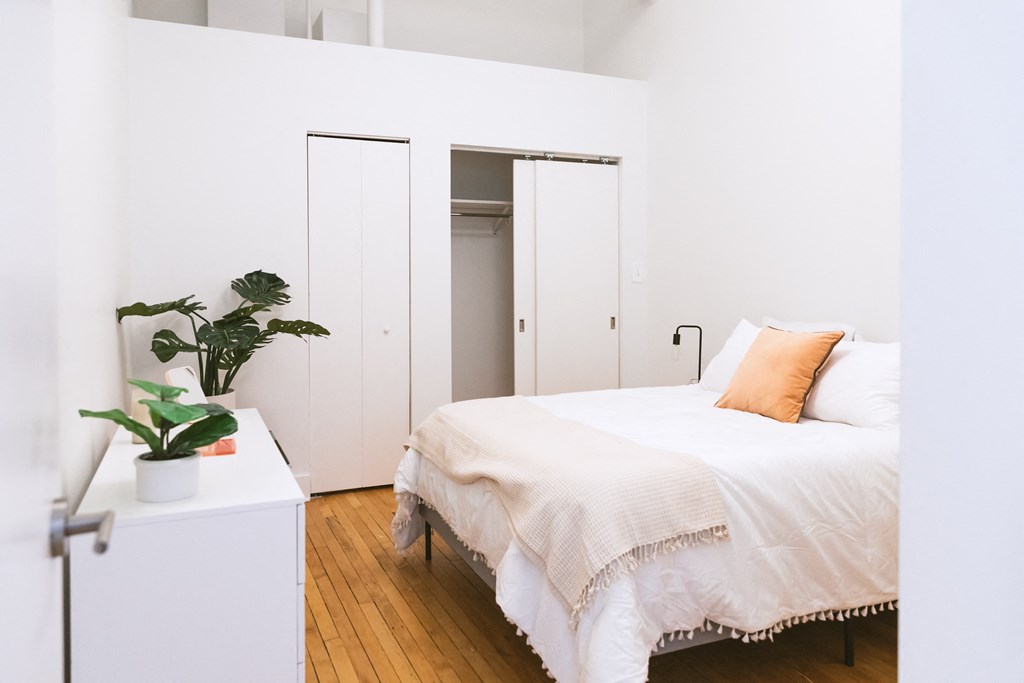 a bedroom with white walls and a white bed with an orange pillow at The Ferguson Apartments, Detroit, 48226