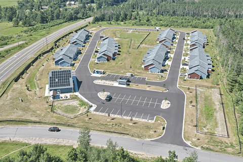 A road with a car driving down it and a parking lot with a building in the background.