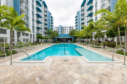 A swimming pool surrounded by palm trees and lounge chairs in front of apartment buildings.
