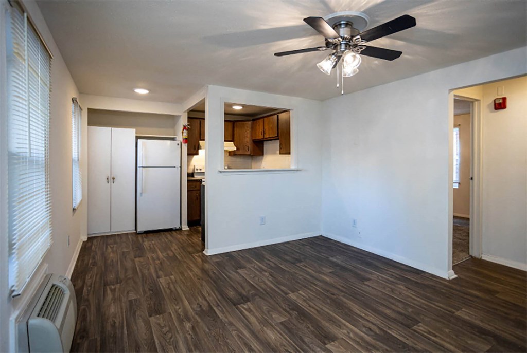 an empty living room with a ceiling fan and a kitchen in the background