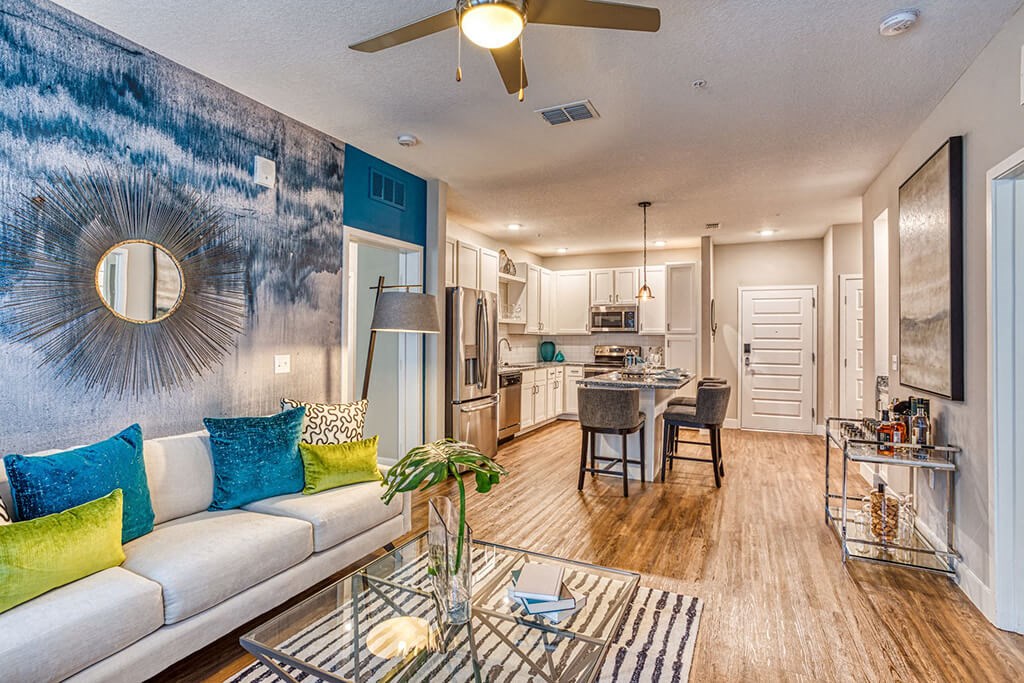 Apartments for Rent in Maitland, FL - The Parker at Maitland Station - Living Room with Wood Vinyl Plank Flooring, Area Rug, Couch, Standing Lamp, Glass-Top Coffee Table, Ceiling Fan, and View of Kitchen.