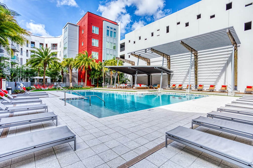 Maitland, FL Apartments - Parker at Maitland Station - Resort-Style Pool with Sundeck, Lounge Chairs, Covered Areas, Umbrellas, Tables and Chairs, Lawn Area with Red Seats, Surrounding Plants and Palm Trees, and View of Apartment Building