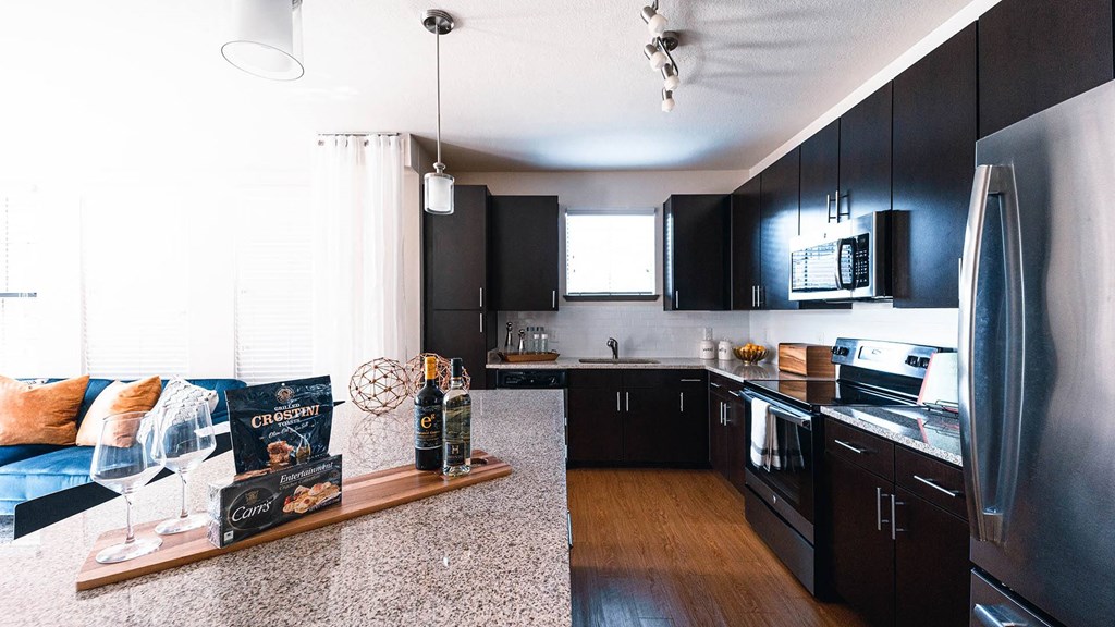 Greenville SC Pet Friendly Apartments - Velo Verdae - Modern Kitchen with Natural Oak-Style Plank Flooring, Stainless Steel Appliances, White tile Backsplash, Granite Countertops, Cooking Island, Dark Wood Cabinetry, and Overhead Lighting