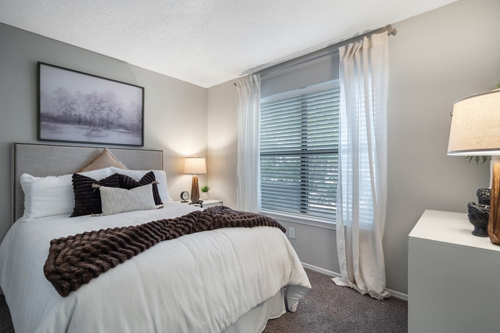 A bedroom with a large bed and a window with white curtains.