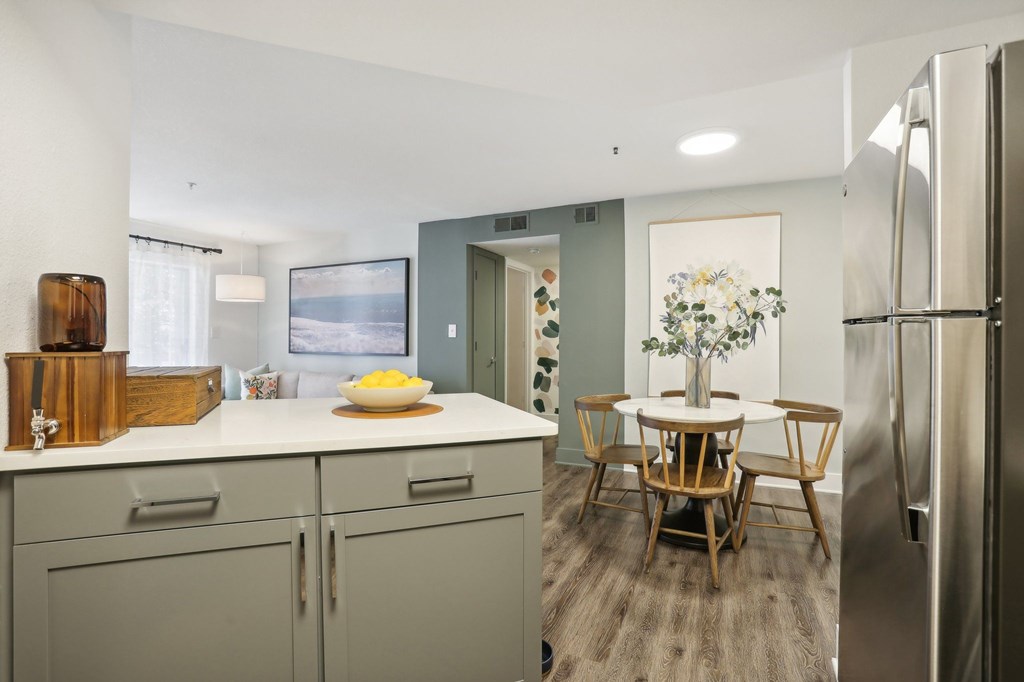 A kitchen with a refrigerator, a dining table with chairs, and a bowl of fruit on the counter.