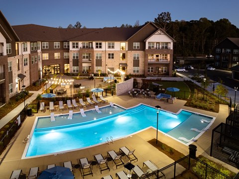 A large swimming pool surrounded by lounge chairs and umbrellas in front of a multi-story apartment building.