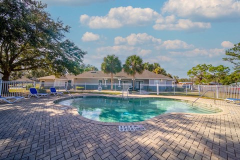 A pool surrounded by a brick patio with a white fence and trees in the background.
