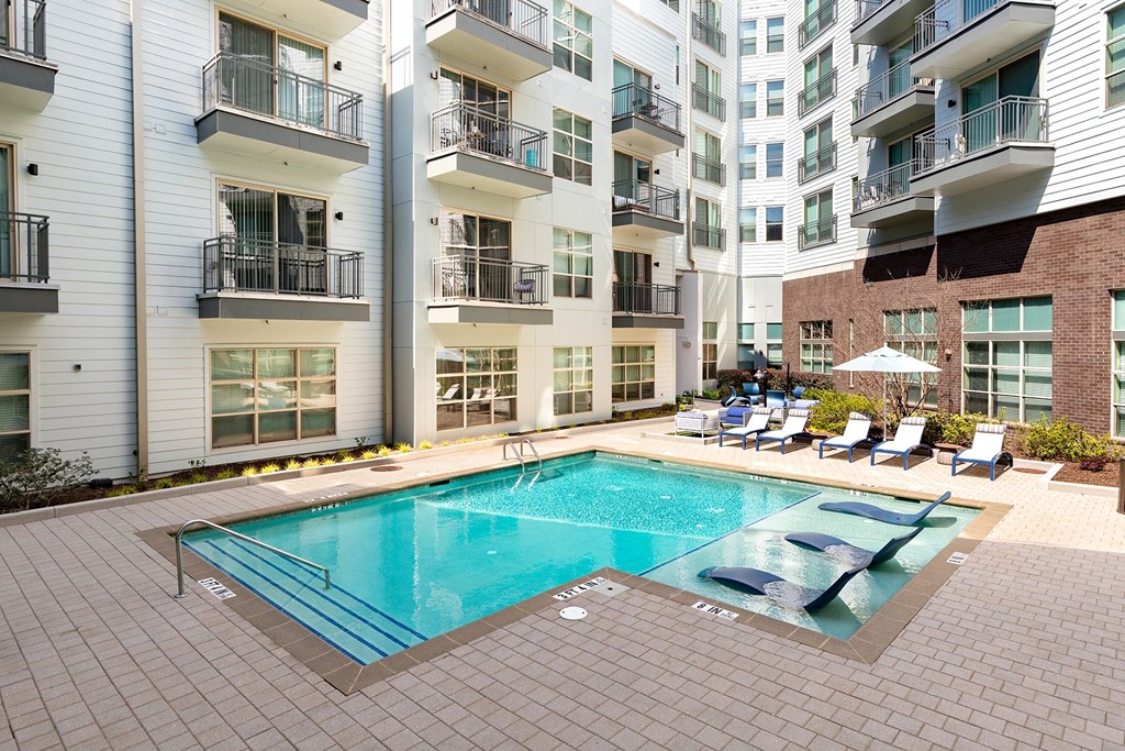 Two-Bedroom Apartments in Decatur, GA - Arlo Decatur - Resort-Style Pool With Poolside and In-Pool Lounge Seats, Umbrella, and View of Apartment Buildings