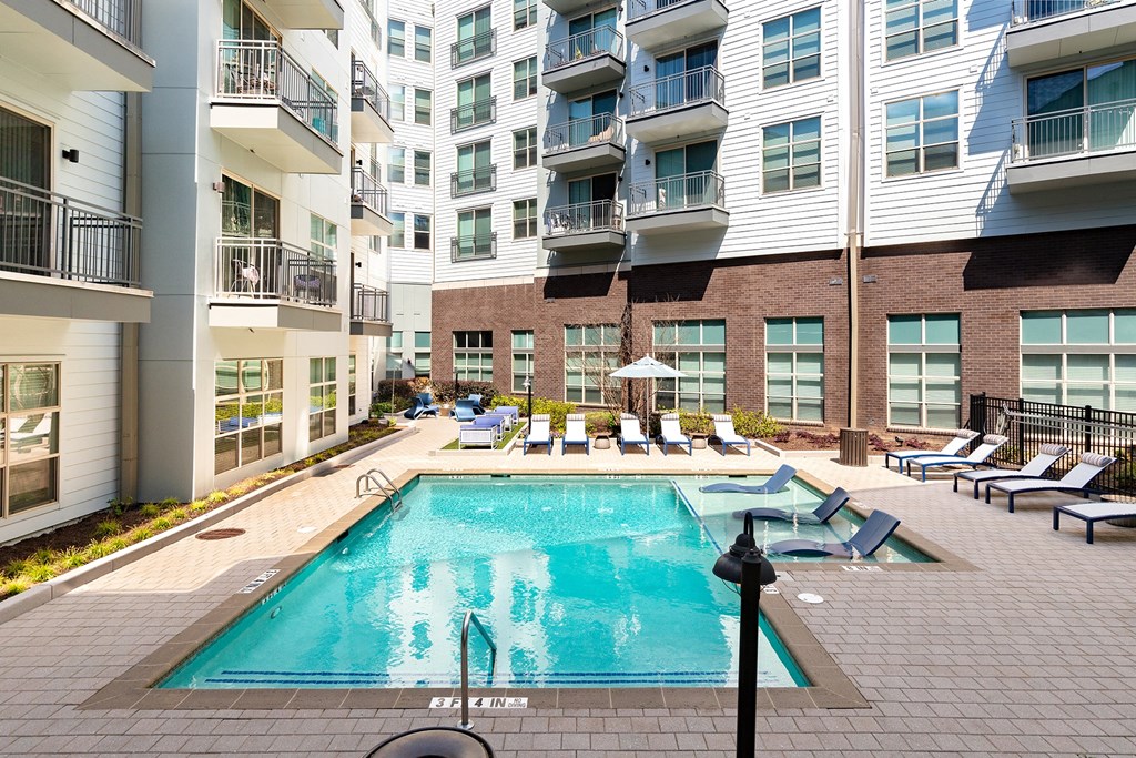Decatur, GA Apartments Near Decatur High School - ARLO - Resort-Style Pool With Spacious Sundeck, Submerged And Poolside Lounge Seats, Umbrellas, And Surrounding Apartment Building.