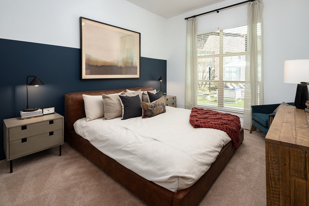 1-Bedroom Apartments in Decatur, GA - Arlo Decatur - Bedroom With Bed, Nightstands with Lamps, Dresser, Armchair, Plush Carpet, Accent Wall,  Framed Wall Art, and Large Window With Blinds and Coverings