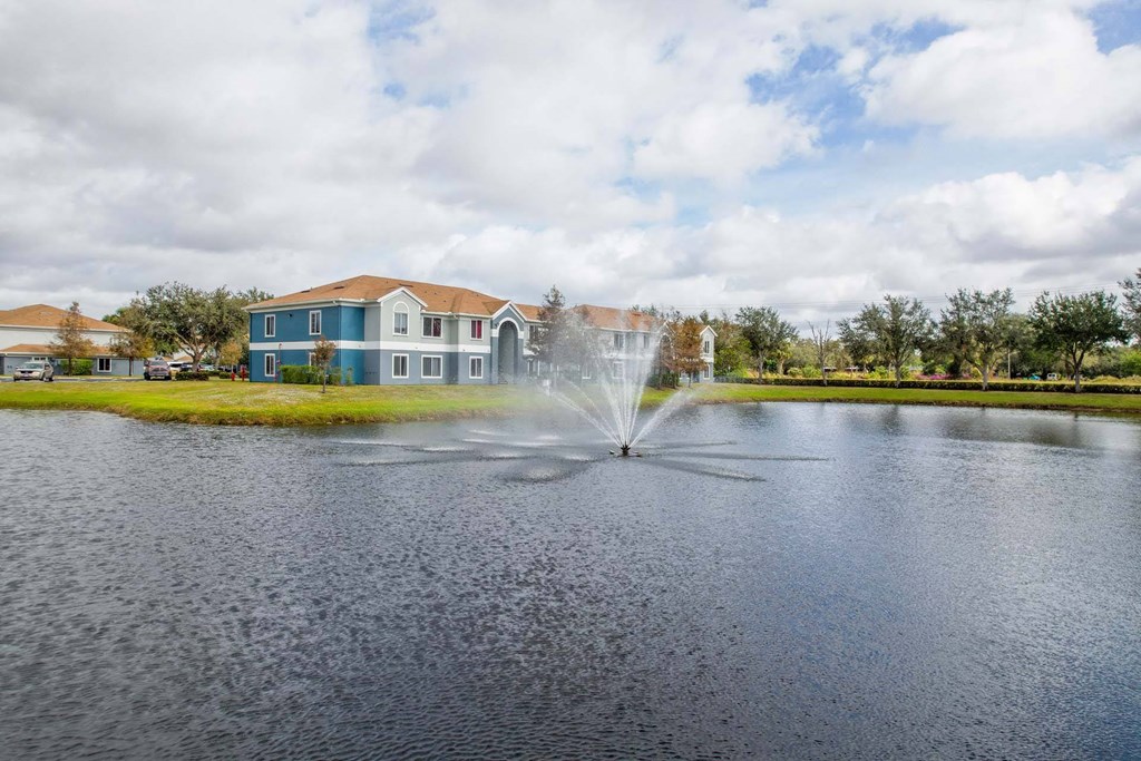 A fountain in the middle of a lake with houses in the background.
